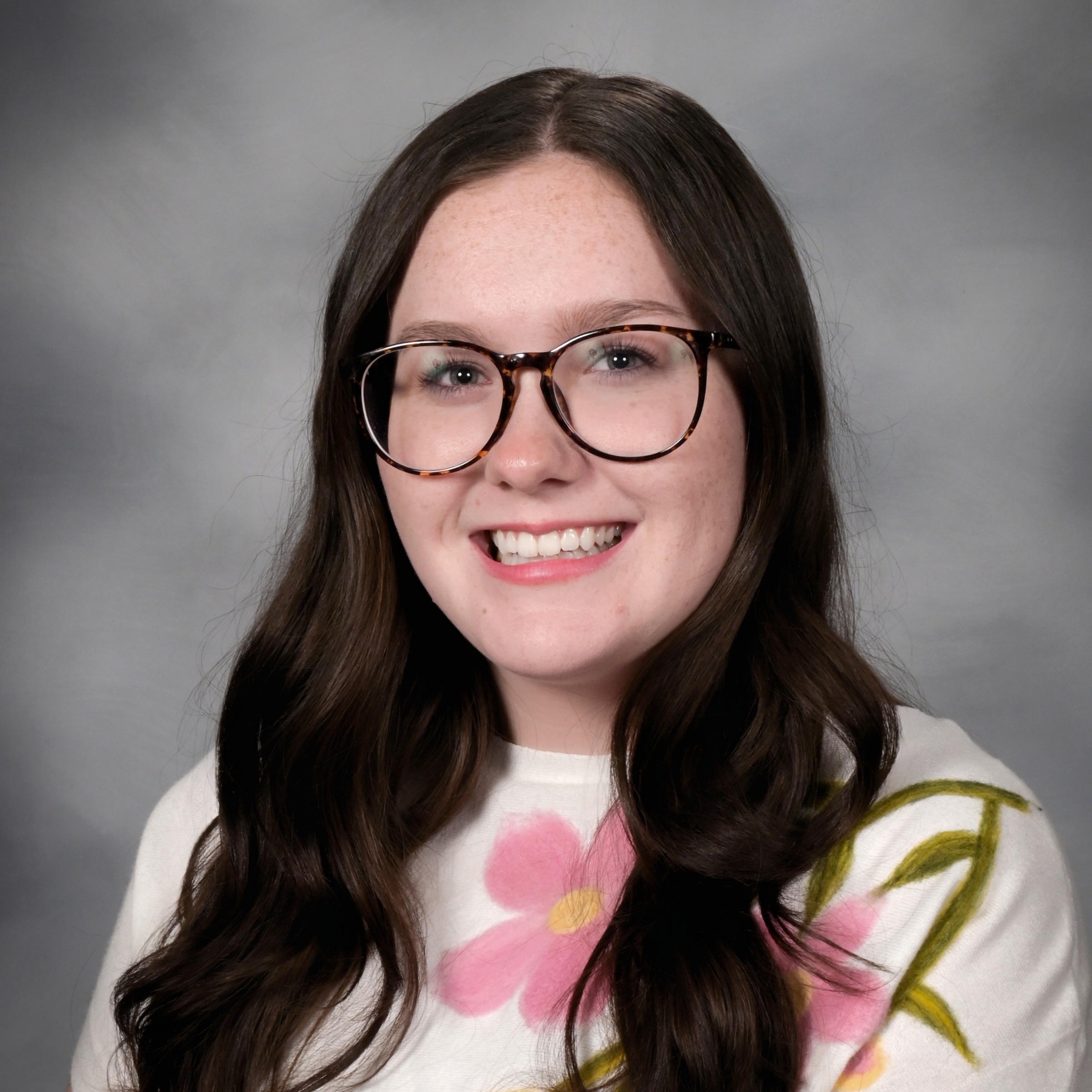 A woman with long brown hair and glasses smiles. She wears a white shirt with a floral print.