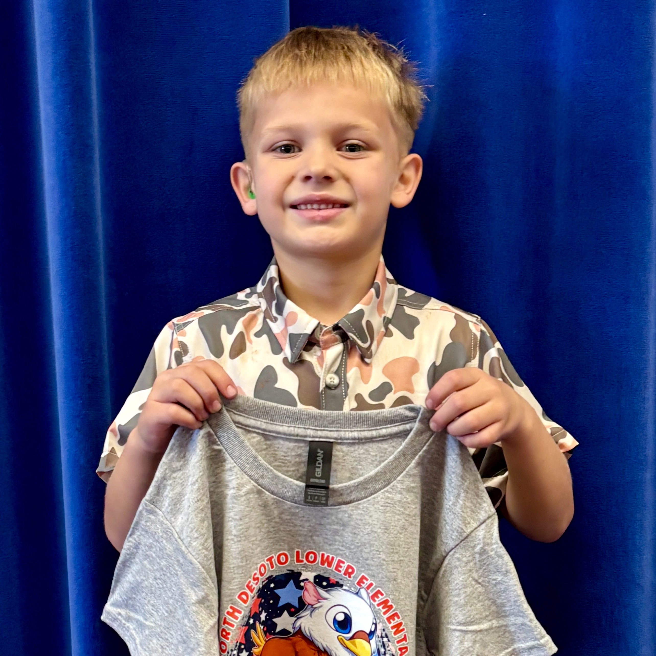 A boy in a camouflage shirt holds up a gray tee with an eagle design. Blue curtains are behind him.