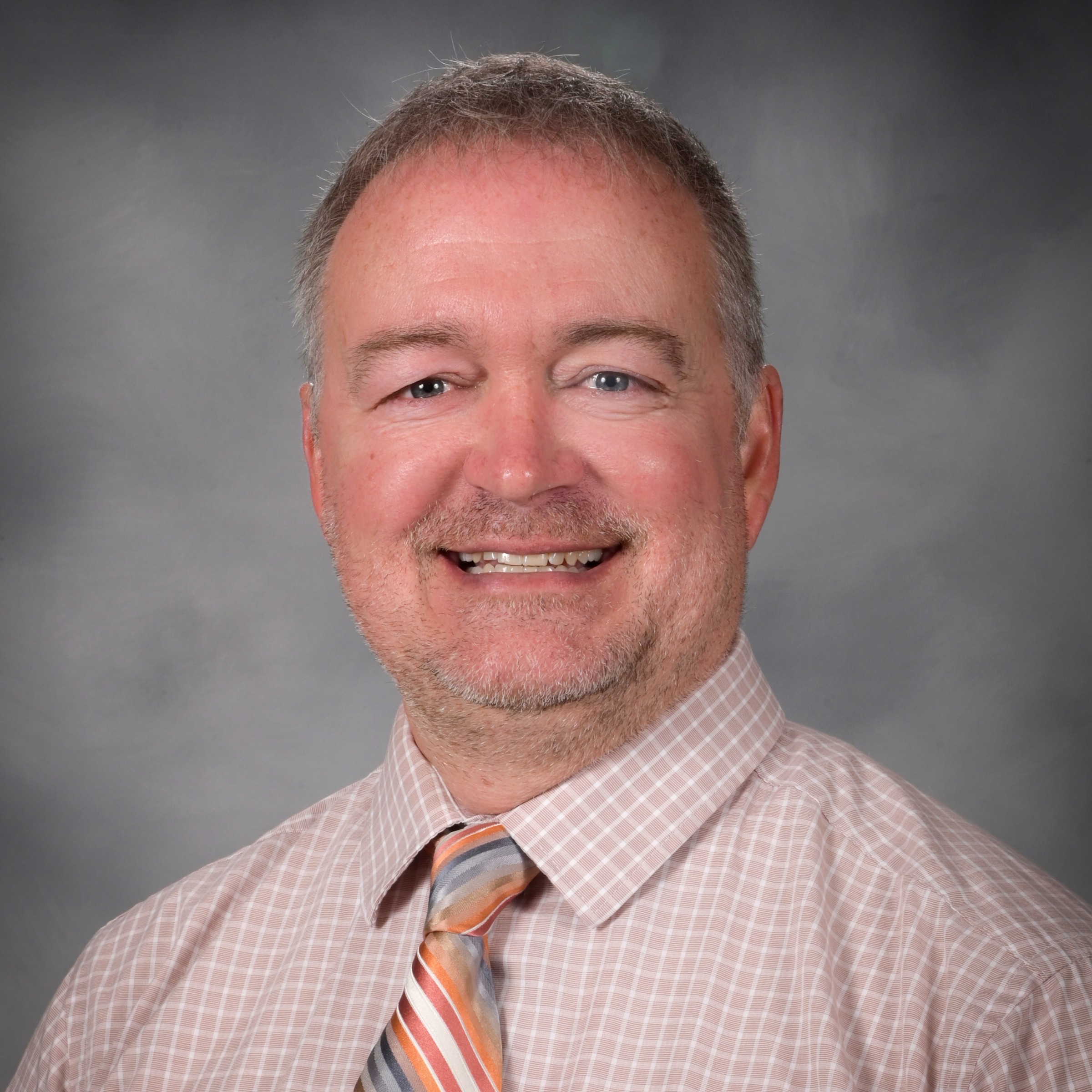 A man with graying hair and a beard, wearing a light pink checkered shirt and a multicolored tie, smiles at the camera against a gray background.