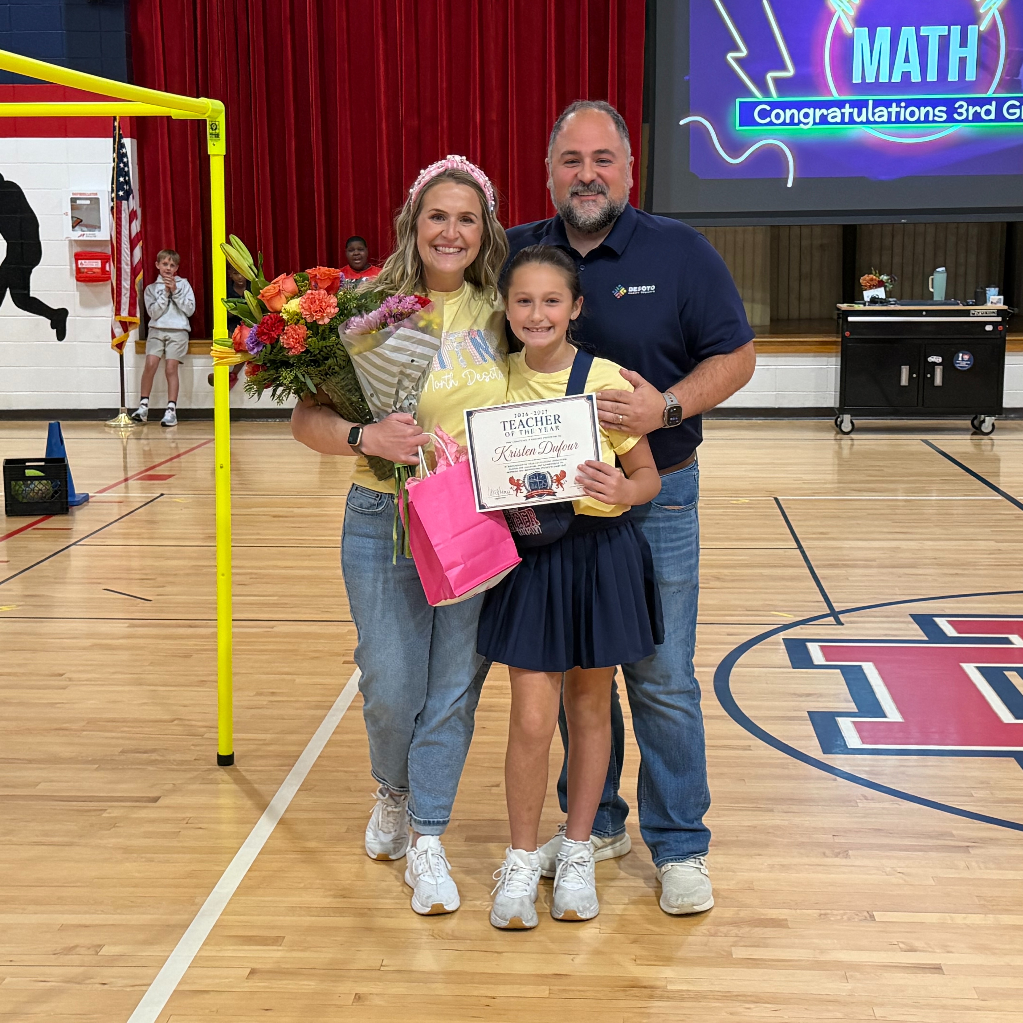 Three people on a basketball court, one holding a certificate, another holding flowers, and a third in a yellow shirt.