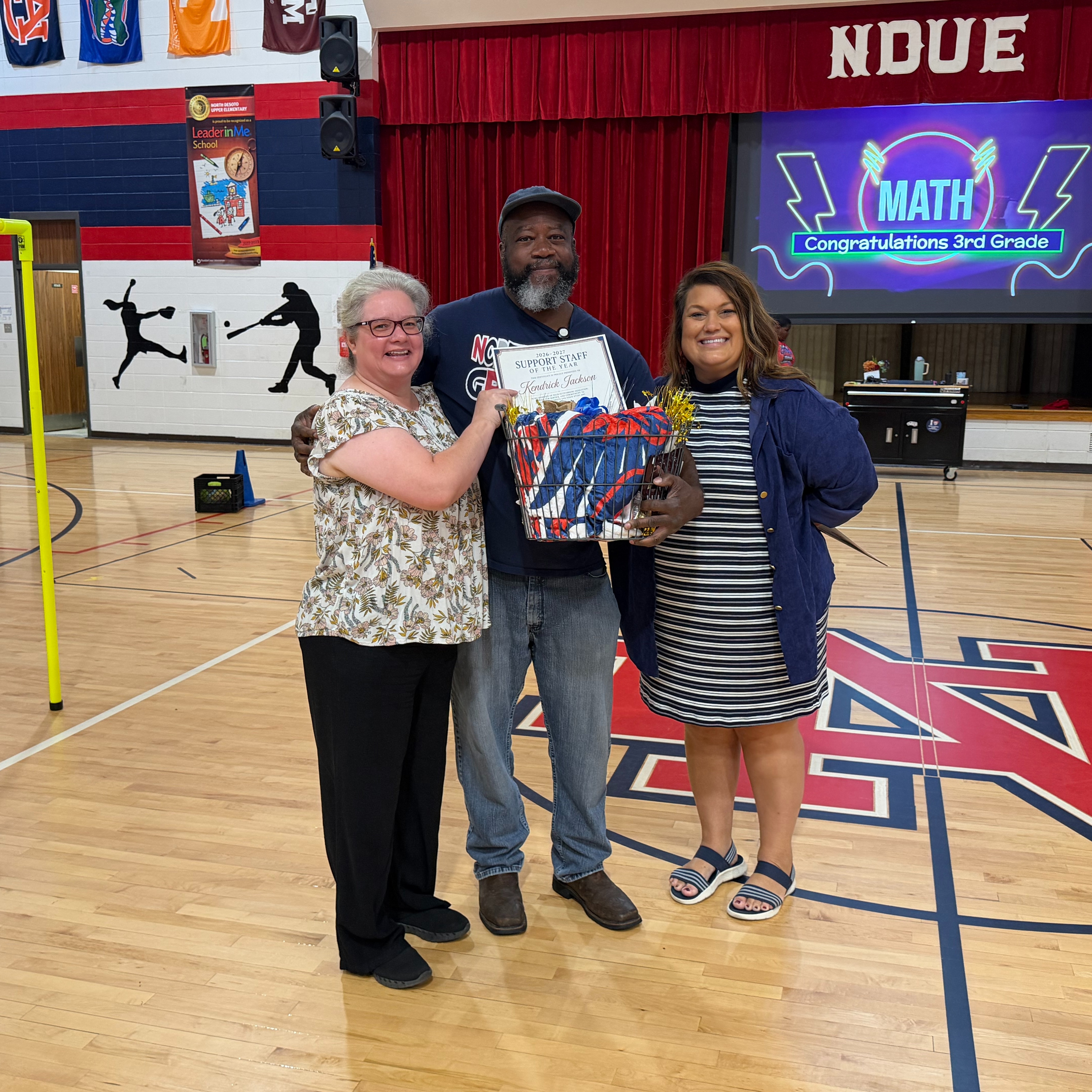 Three people stand in a gymnasium, one holding a certificate. A digital screen reads "MATH". Behind them is a red curtain.