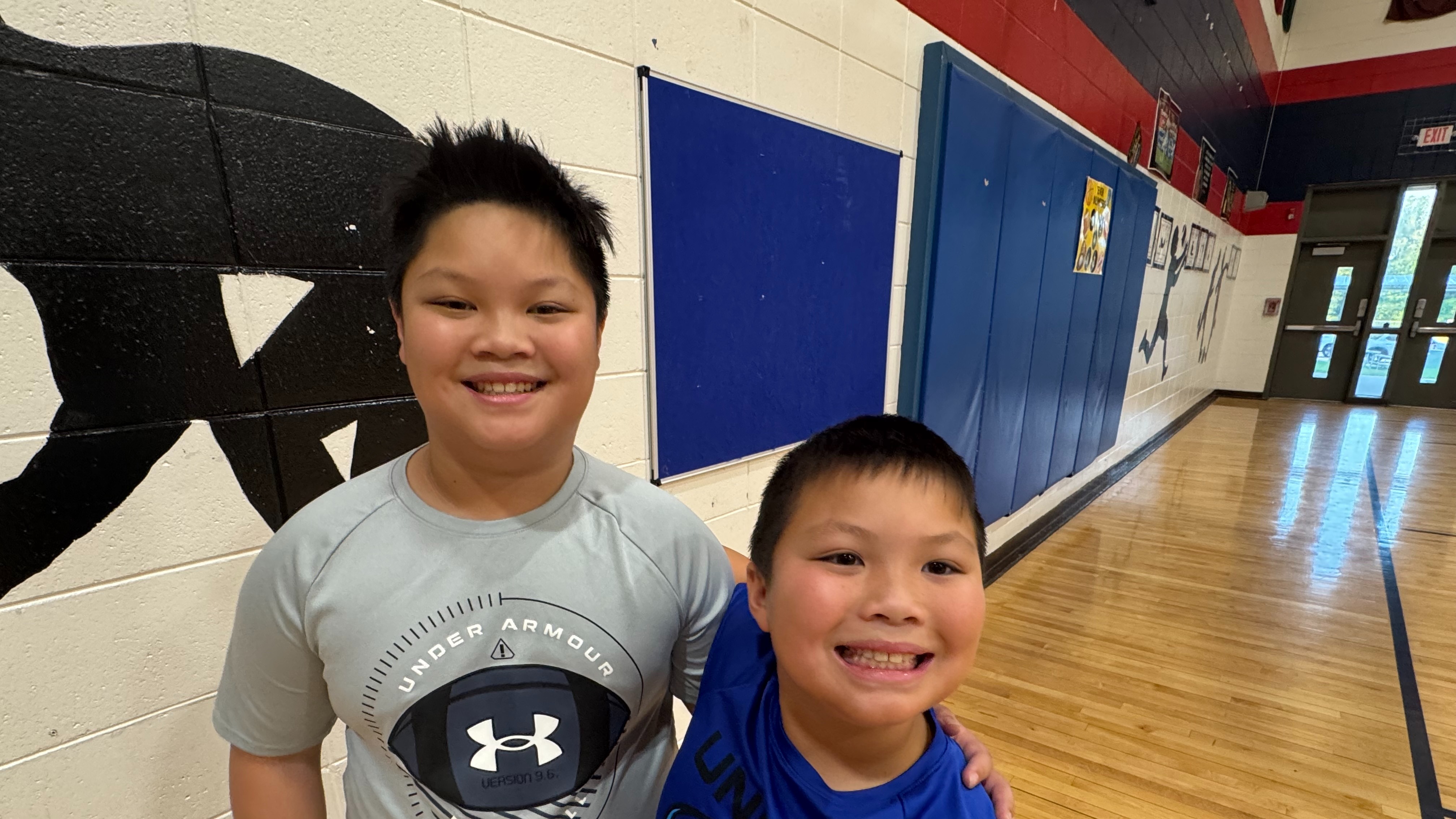 Two boys, one wearing a gray Under Armour shirt and the other in a blue shirt, stand together in a school gym.