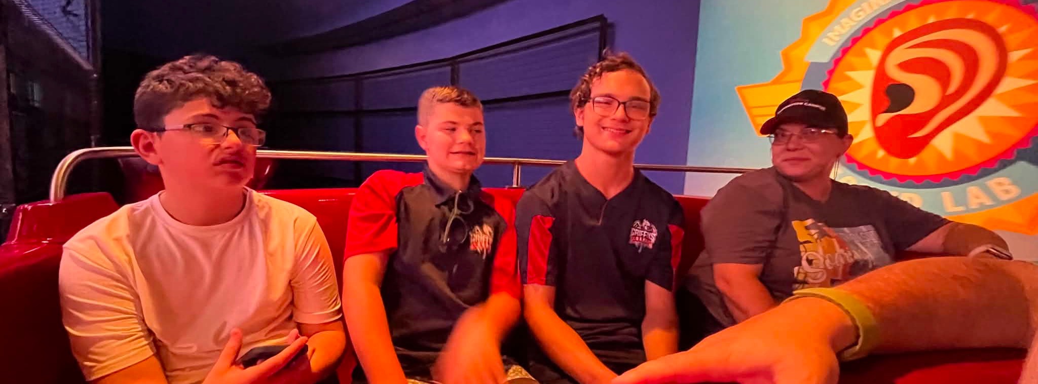 Four boys sit in a ride vehicle with a red seat. Three wear glasses. Behind them is a large screen.