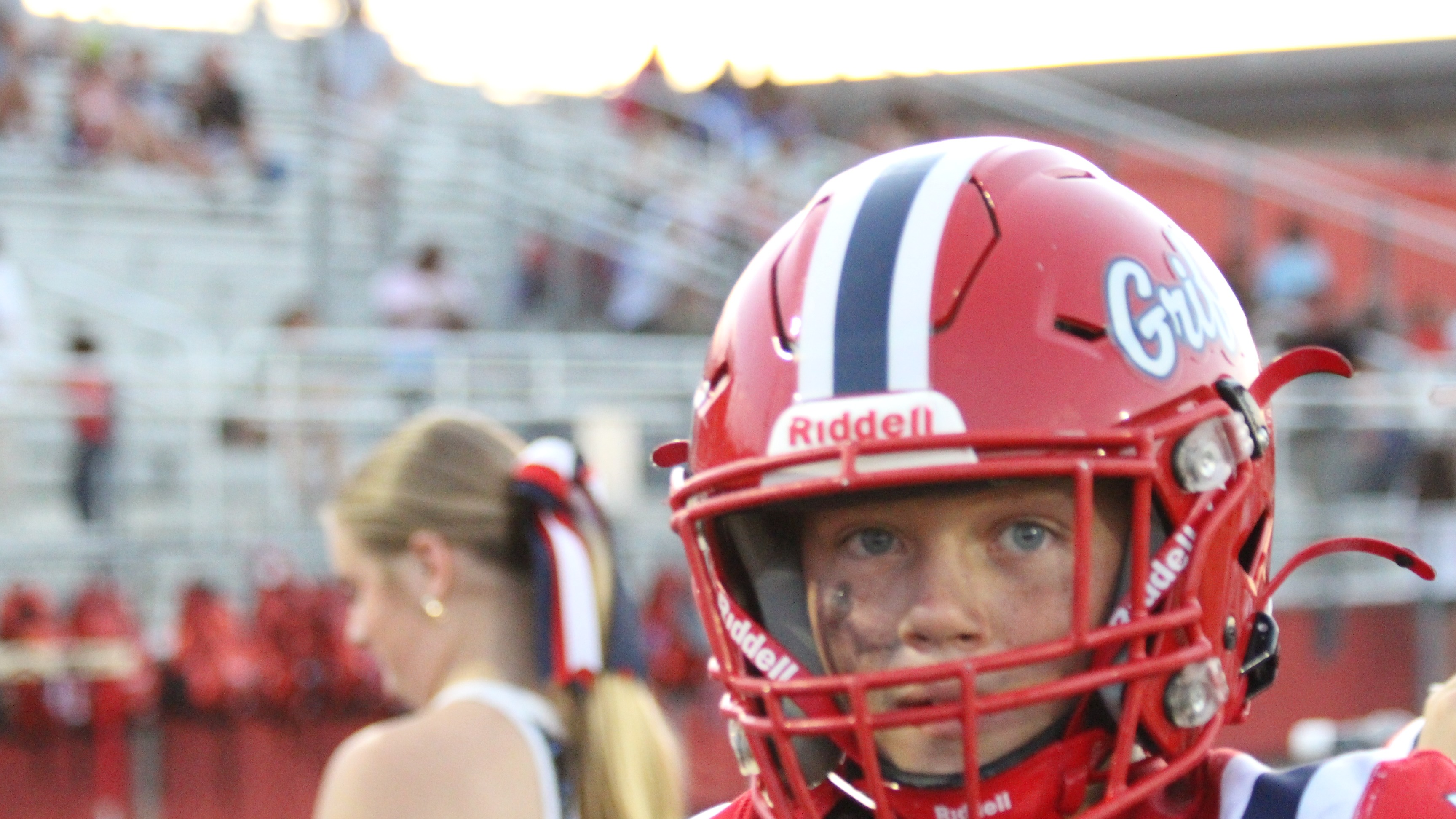 A person with a red helmet labeled "Grit" stands in front of a blurred person and stadium seating.