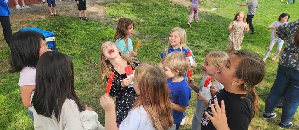 Children on a grassy field, some holding and eating ice pops. Some wear shoes and sneakers.