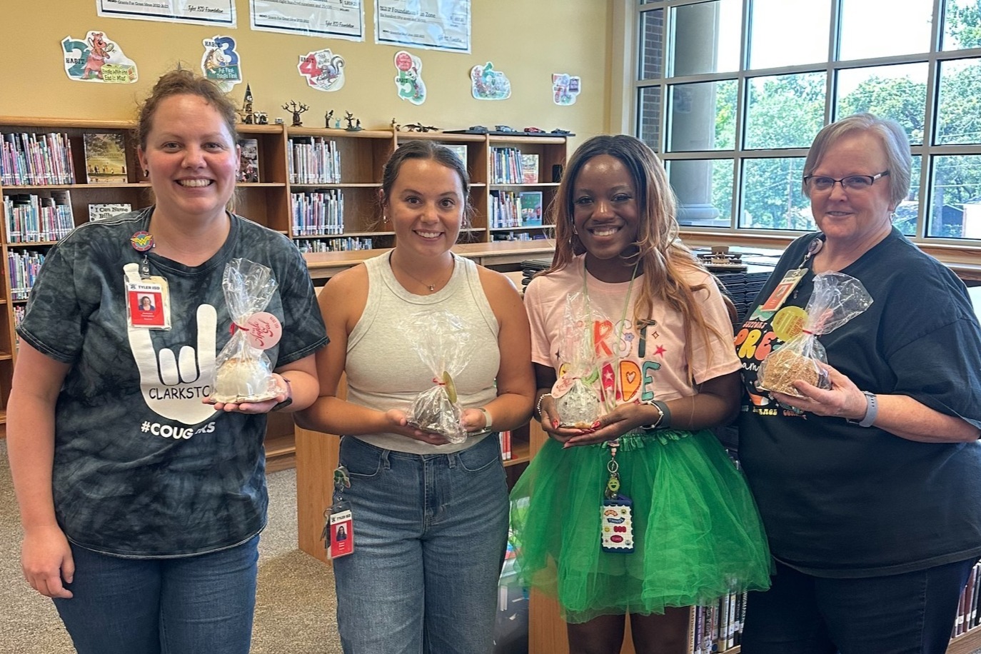 Four women in a library pose for a photo, holding items in their hands. Behind them are bookshelves and windows.