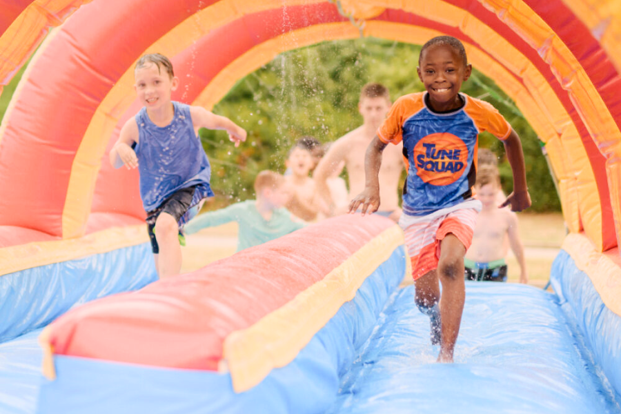an African American student and a Caucasian student both wearing swim clothes running on a red, blue, and yellow inflatable water slide.