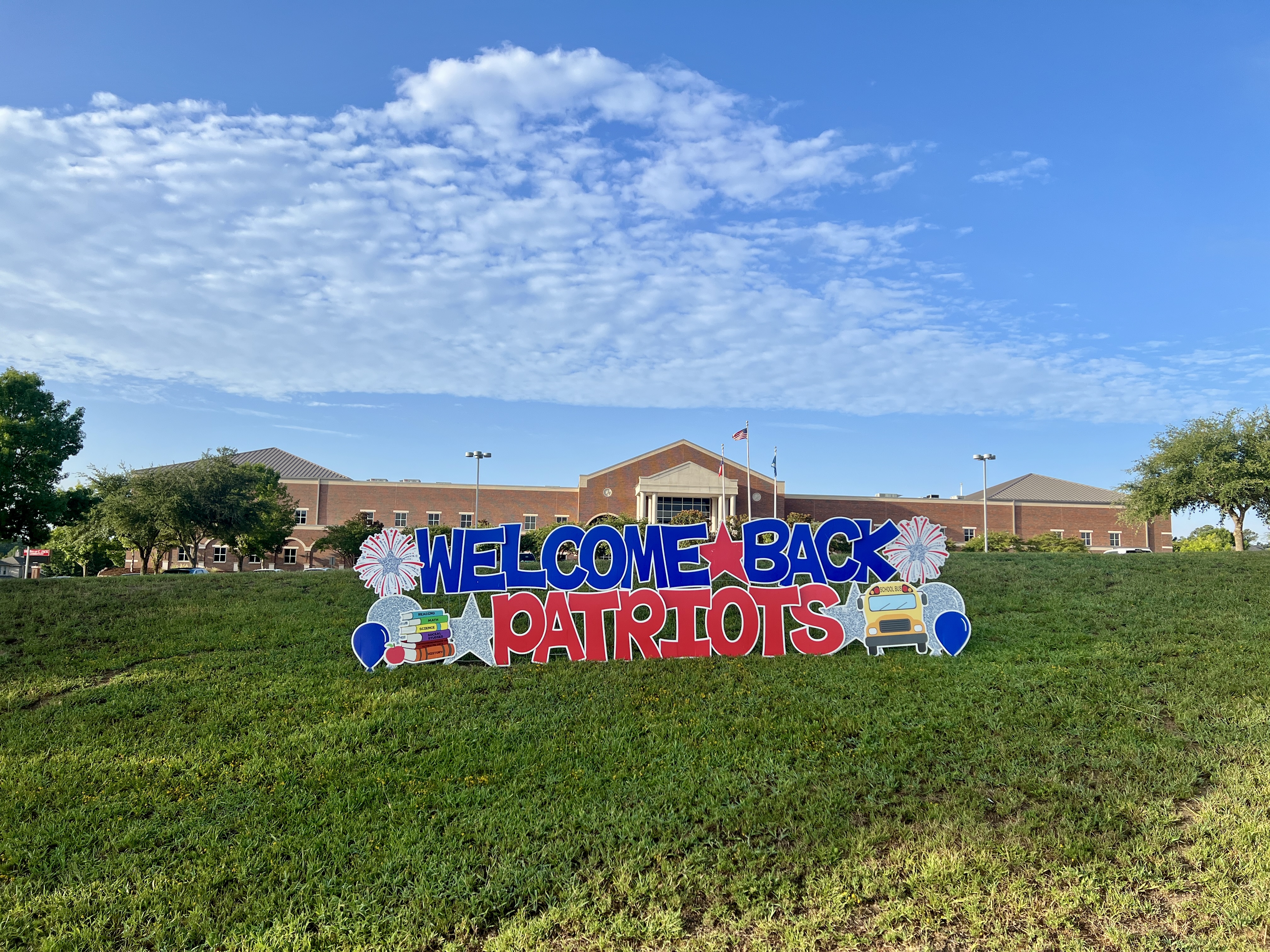 Large, colorful sign on a grassy field says "Welcome Back Patriots" with balloons. Behind is a building under a partly cloudy sky.