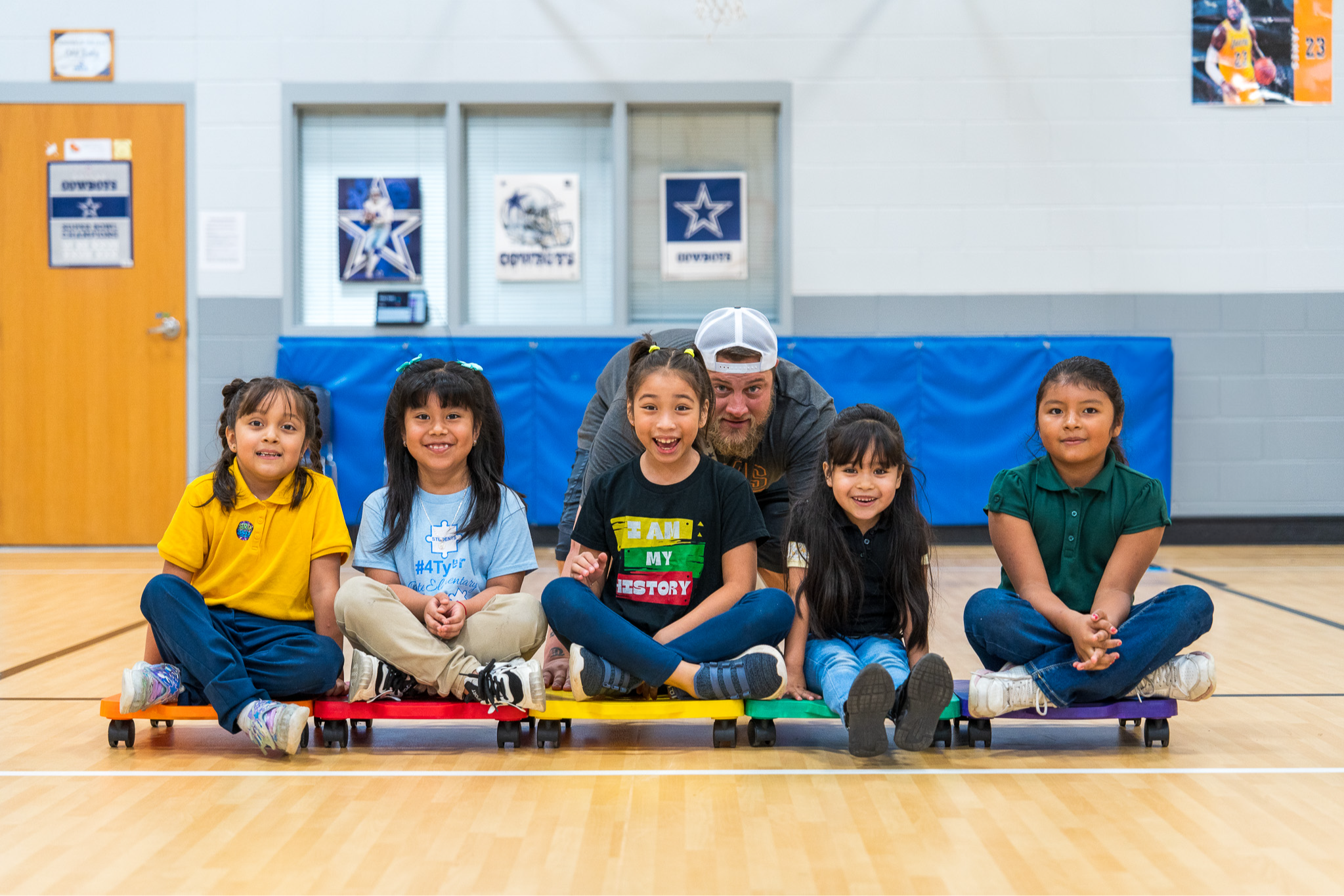 man pushing five elementary age kids on scooters in a gym