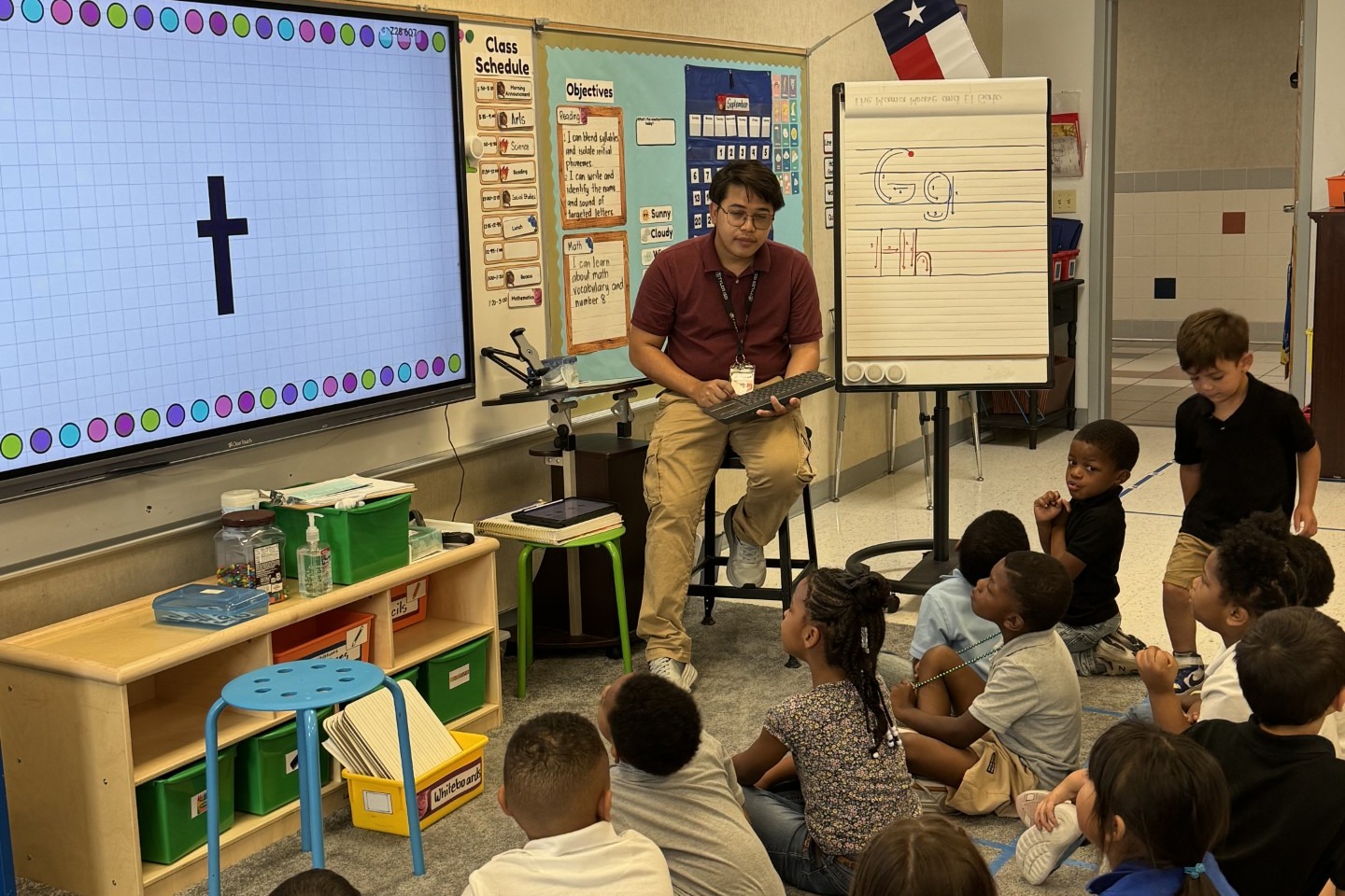 A classroom with children seated on the floor. A teacher is sitting on a stool, holding a tablet.