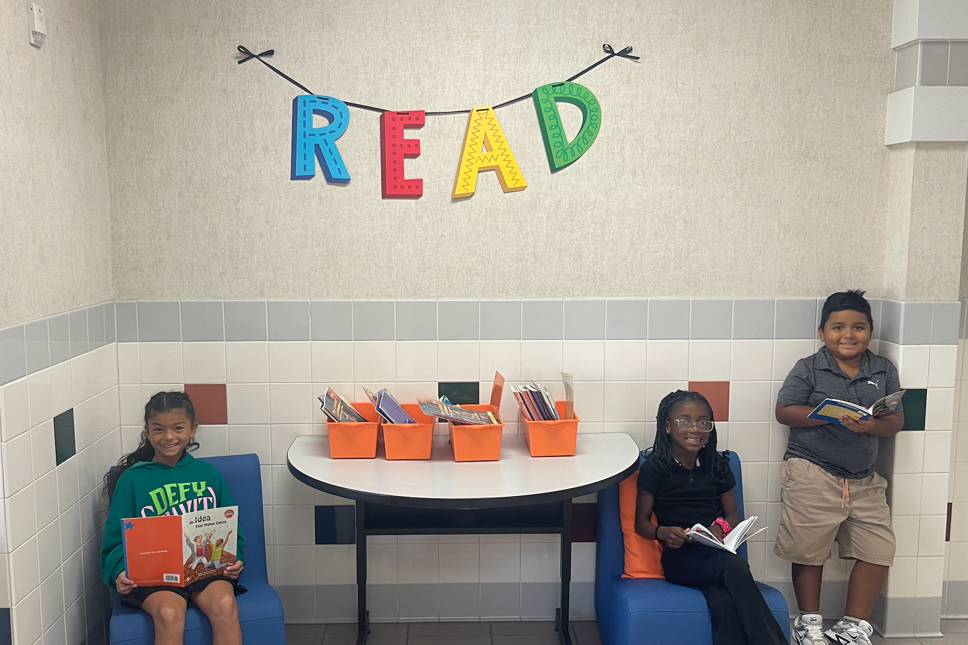 Three children seated at a table in a room with a colorful "READ" banner on the wall.