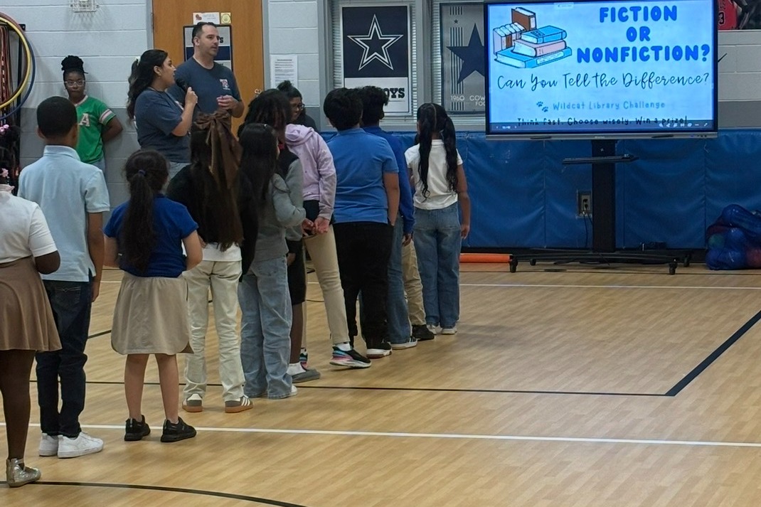 A group of children stands in a line in a gymnasium, facing a large screen displaying "Fiction or Nonfiction?"