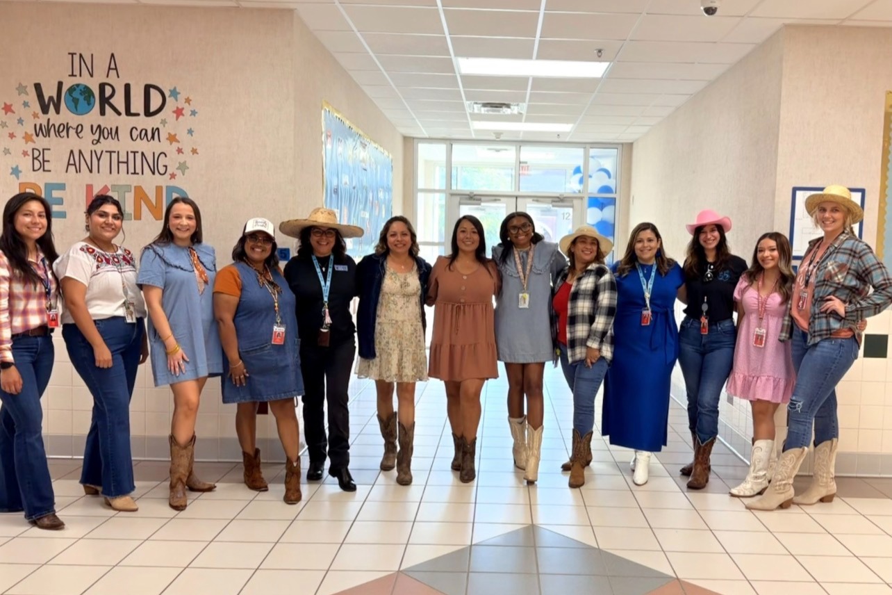 A group of women in various dresses and boots stand in a hallway, smiling, with an inspirational wall behind them.