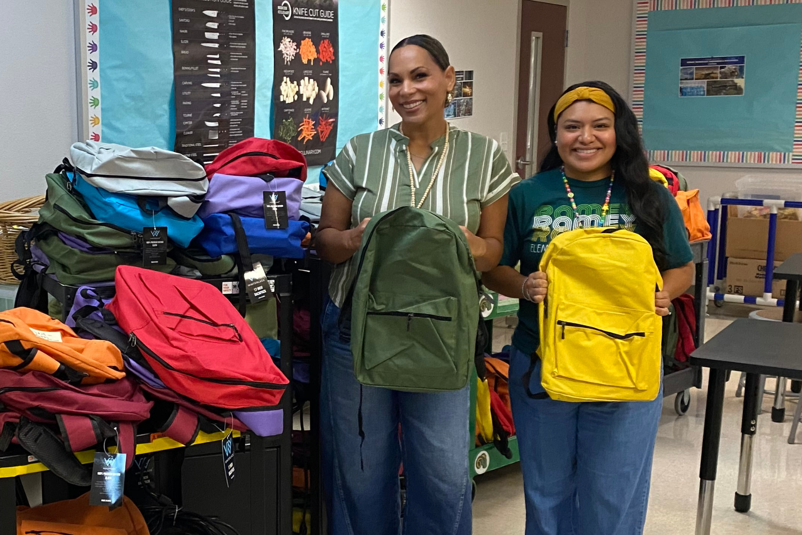 Two women stand next to backpacks in a classroom. One holds a green backpack. Piles of colorful backpacks are behind them.