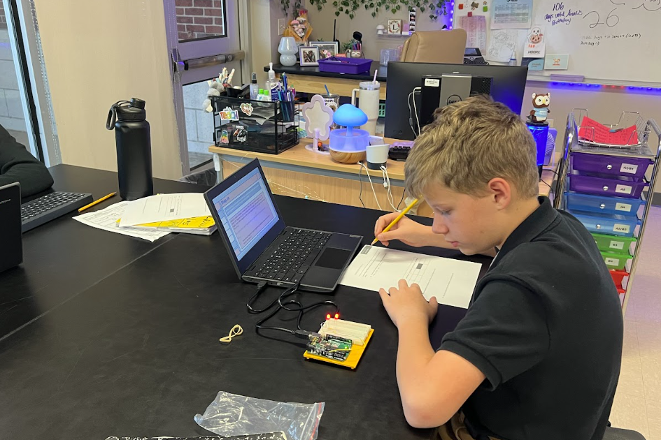 A student writes on paper, focused on a laptop on a desk, with various tech equipment and a whiteboard in the background.