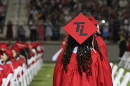 A person in a red graduation gown walks in a line, facing the crowd in a stadium.