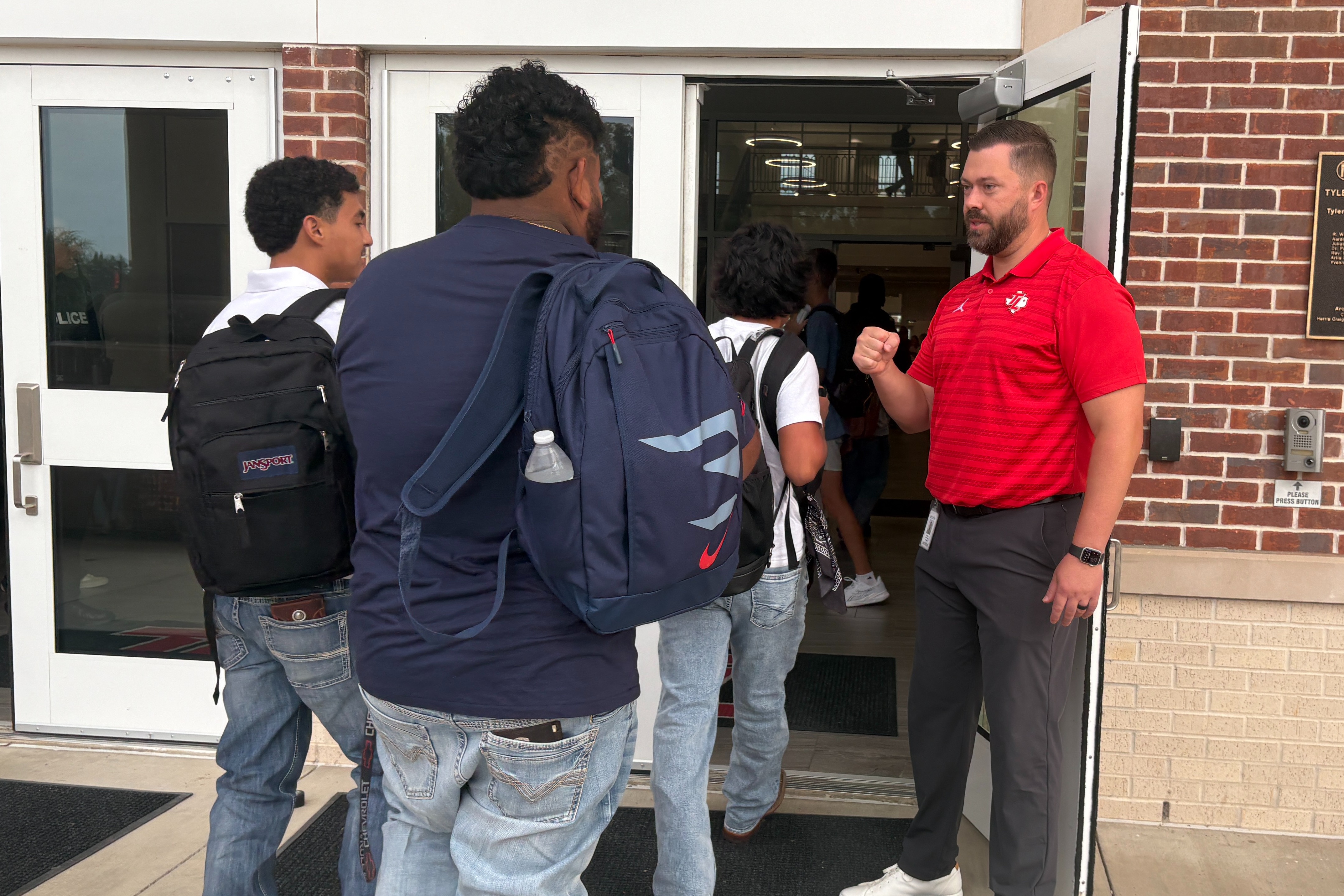 Several people stand in front of a brick building with glass doors. A man in a red shirt greets a student.