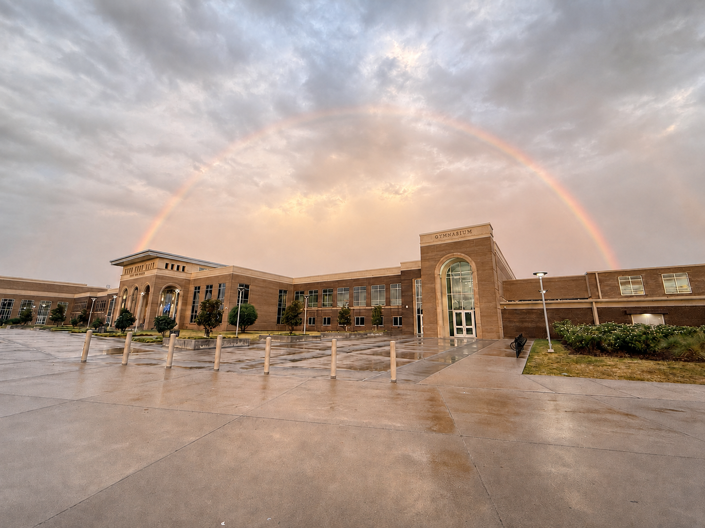Building with rainbow in sky. Large building with multiple windows. Rainbow spanning over building. Wet pavement in foreground.