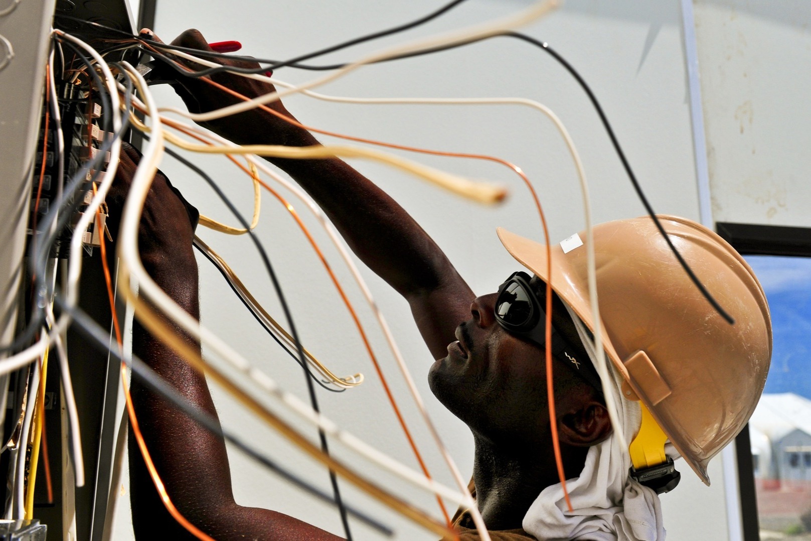 A student works on electrical wiring with wires around him; he wears a yellow hard hat and sunglasses.