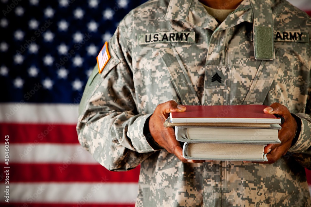 Student wearing United States Army fatigues carrying books in front of an American flag.