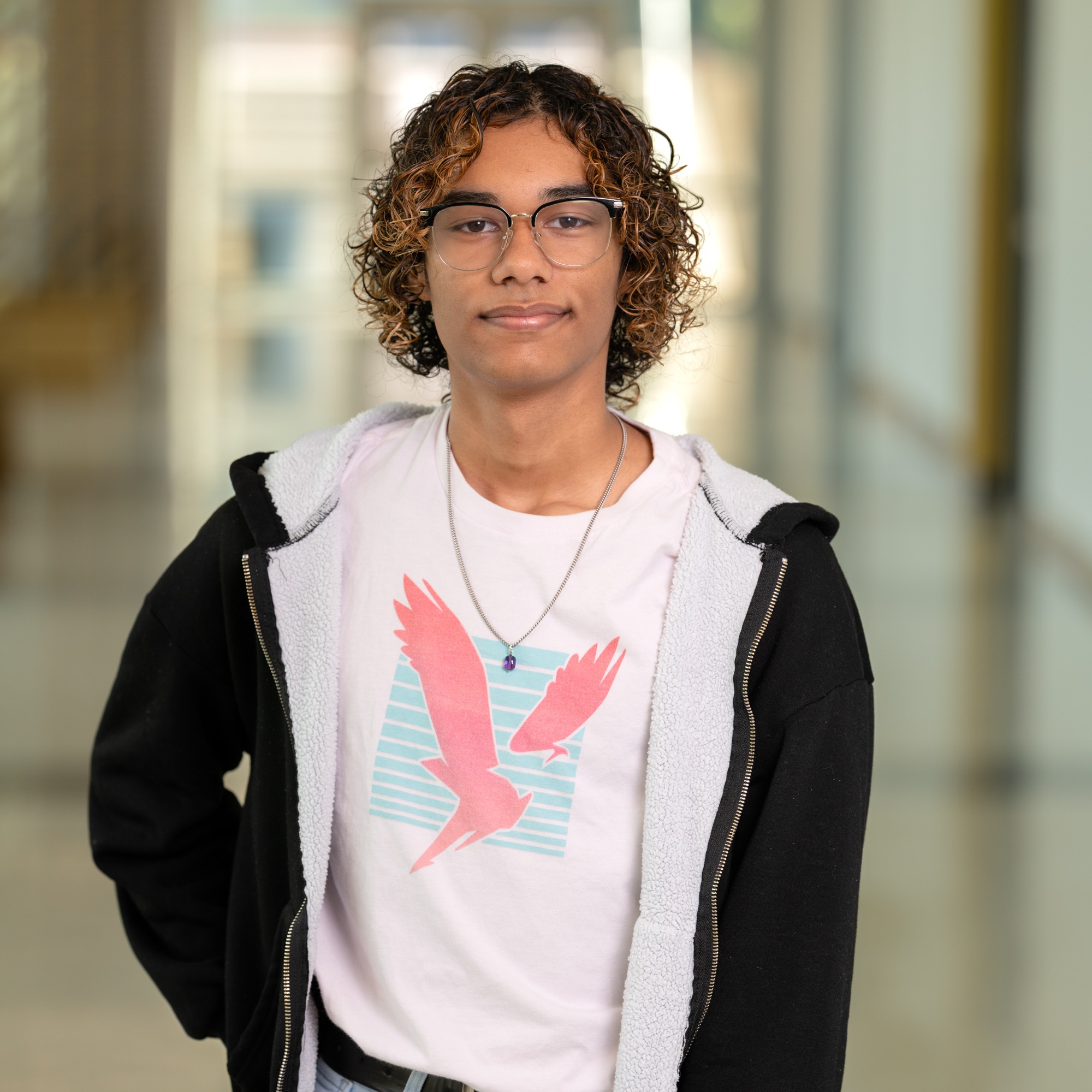 teen boy with curly hair wearing a t-shirt and black jacket standing in hallway