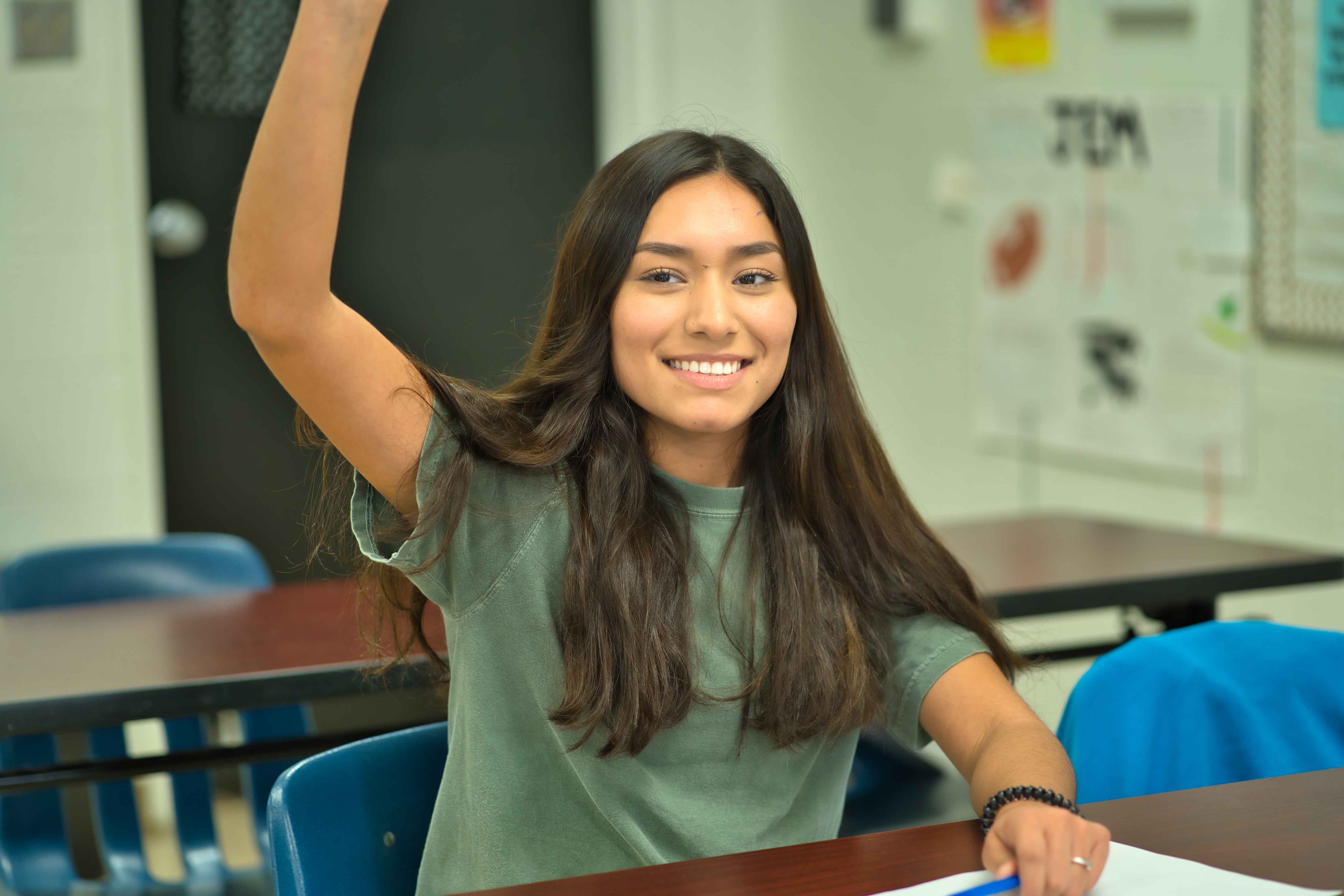 high school female student with long dark hair sitting at a desk with her hand raised.