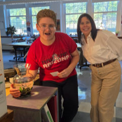 Two individuals in a classroom. A person in a red shirt holding a fish tank, another person in a white shirt, and a beige pants.