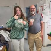 A woman in green and a man in gray stand in a classroom, both holding cards and smiling.
