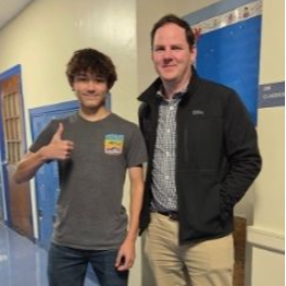 Two men in an indoor hallway. One gives a thumbs up and wears a gray t-shirt. The other wears a black jacket, khaki pants, and sneakers.