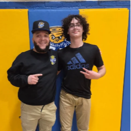 Two men stand in front of a blue and yellow wall. One wears a hat, while the other wears glasses.