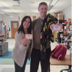 Two people stand in a classroom, smiling. A woman holds a card. A man holds a blanket and a card.