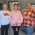Three women stand in a room. One wears a pink hoodie, another a white hoodie, and the third a pink plaid shirt.