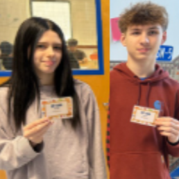 Two students hold badges. The left student has long dark hair and wears a gray sweatshirt. The right student has brown hair and a brown hoodie.