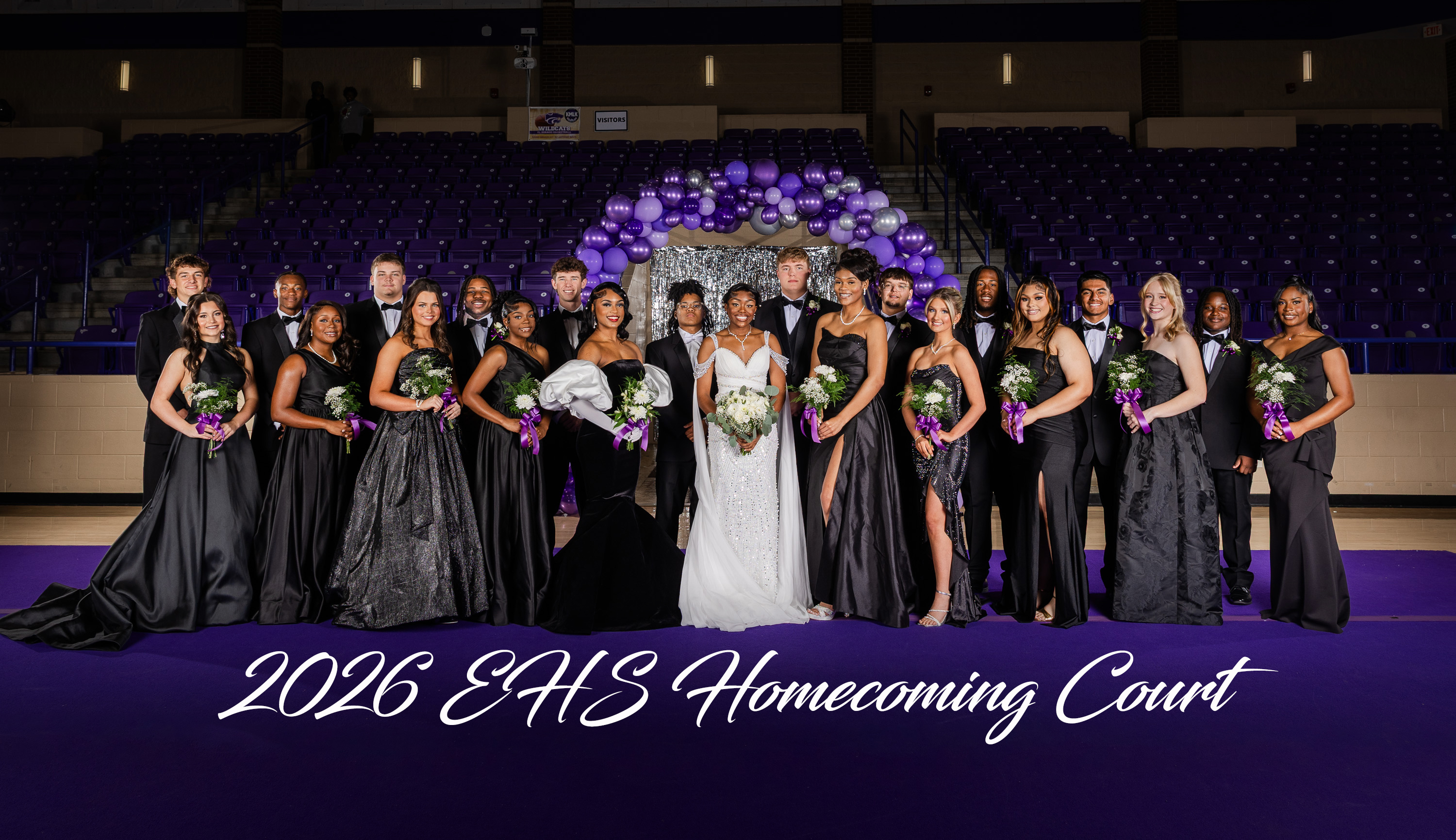 Students in formal wear stand in a gym. A woman in a white gown stands at center.