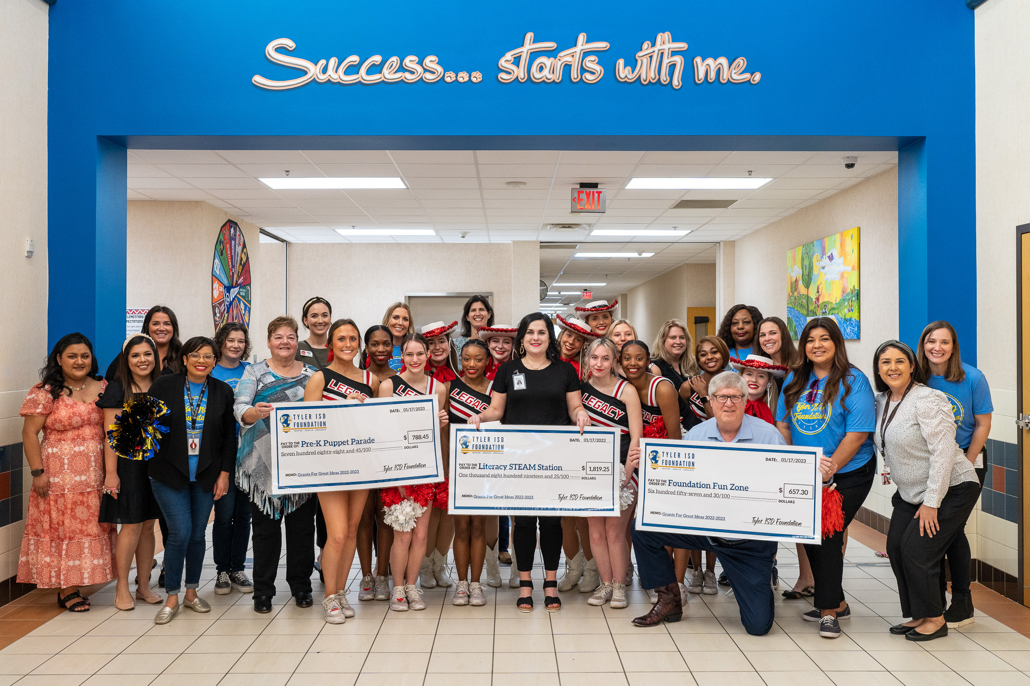 adults standing in a hallway with cheerleaders holding giant checks for grants