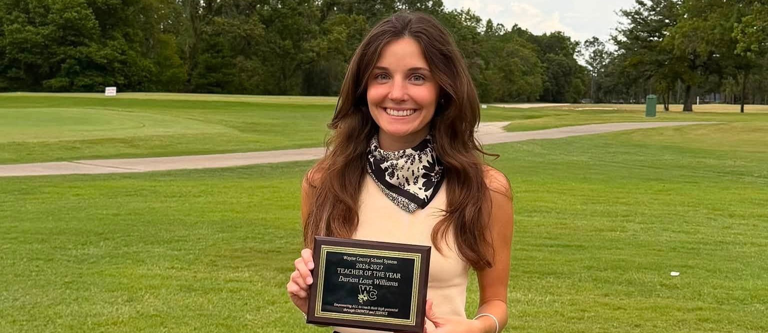 A woman in a black skirt and white top stands on a golf course, holding a plaque.
