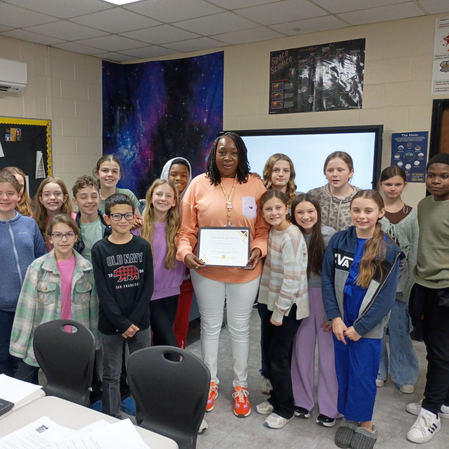 Students gather in a classroom behind a teacher holding an award certificate. They are standing in front of a monitor.