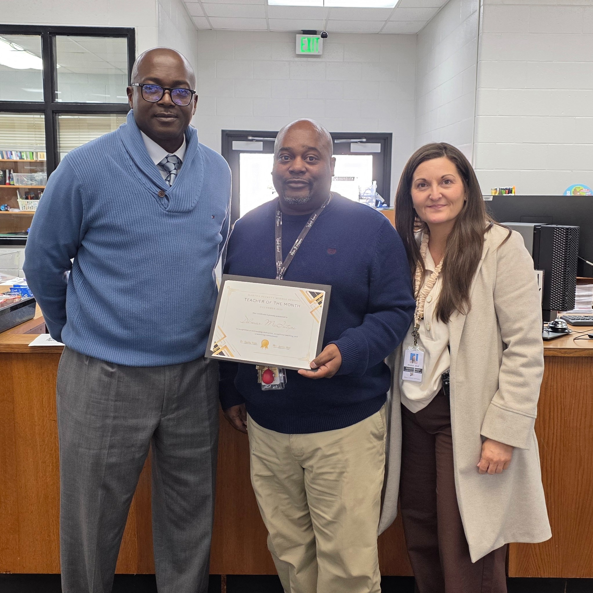 Three individuals stand in a room with a desk. The man in the middle holds a certificate. Behind them is a desk with books and a monitor.
