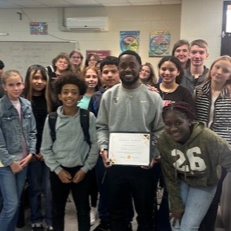 Group of students and two adults posing for a photo in a classroom. One adult holds a framed award.