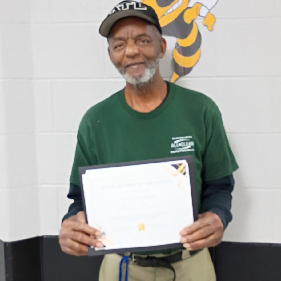 A man in a green shirt and cap stands in a hallway, holding a framed certificate. Behind him is a white wall with a bee emblem.