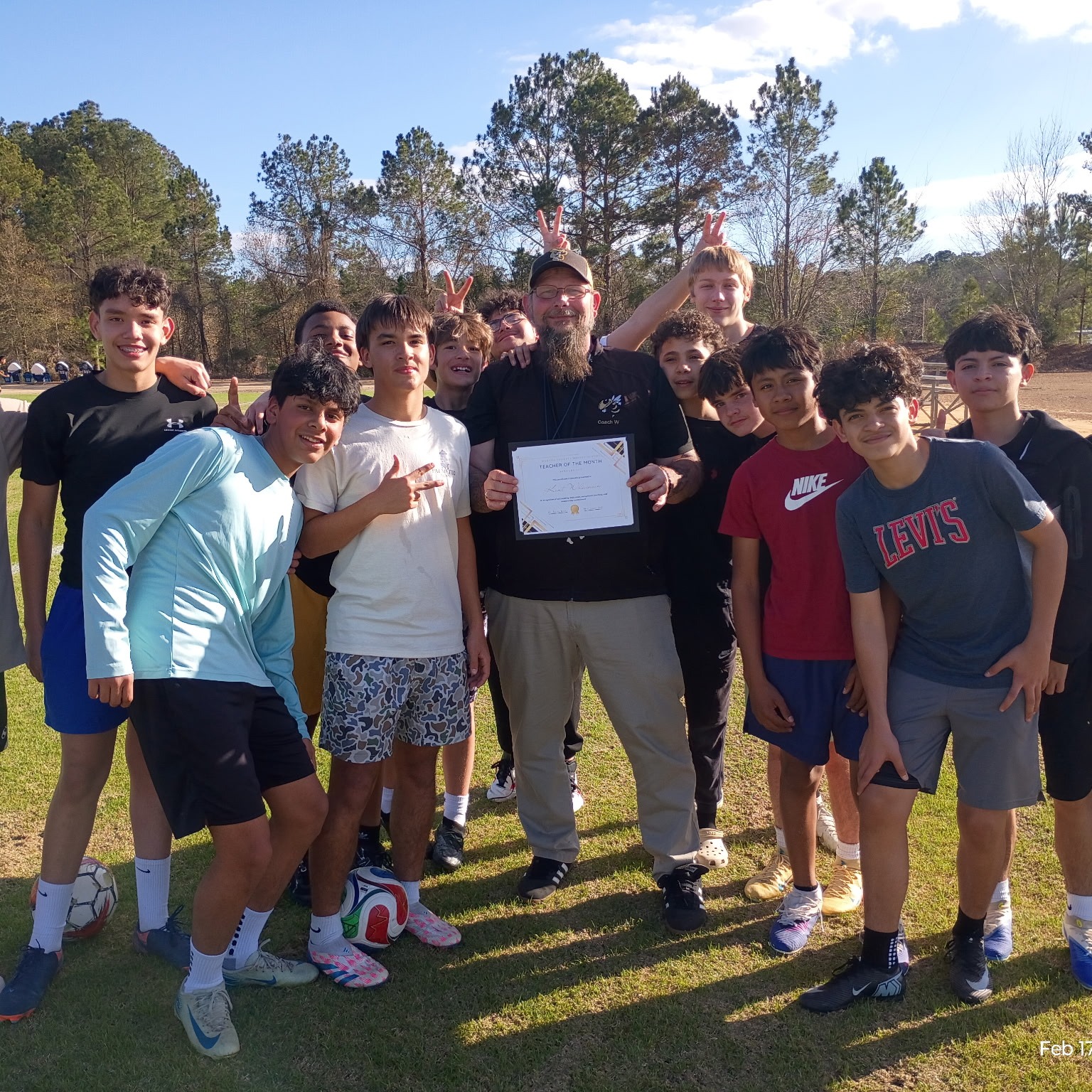 A group of individuals, possibly a sports team, standing on a grassy field. Some wear sports uniforms, sneakers, and hats. A man holds a certificate.