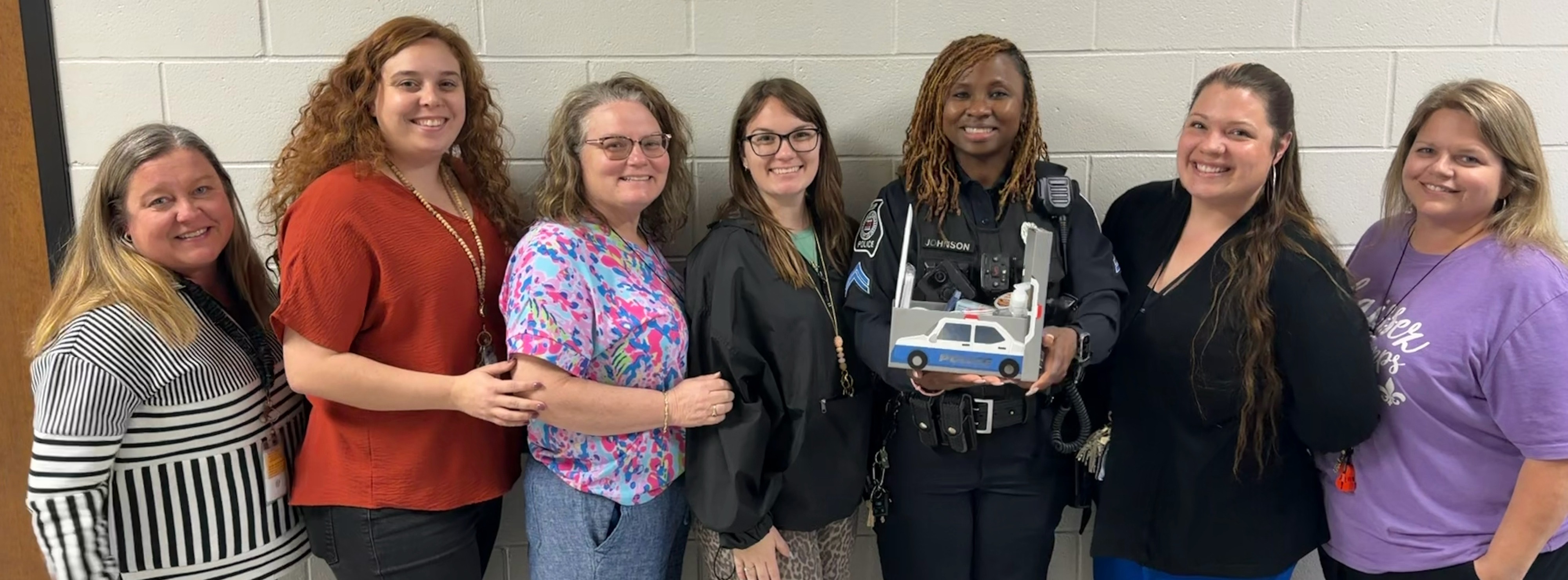 Seven women pose in front of a white wall with a bee logo. They wear various colored tops and pants.