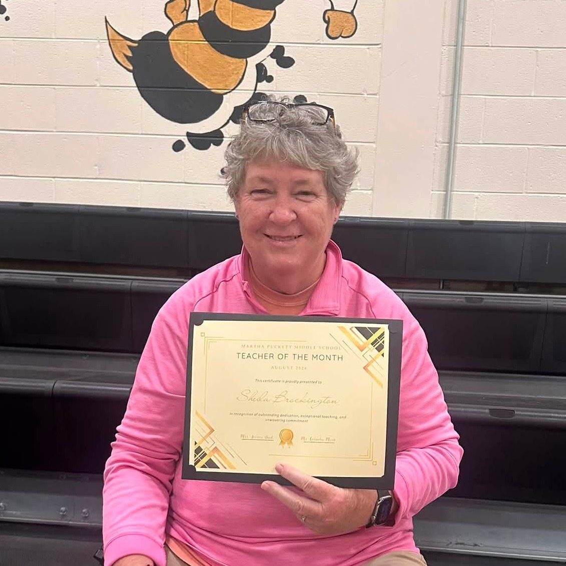 A woman with a certificate sits on a stool, smiling, with a bee logo behind her on a wall.