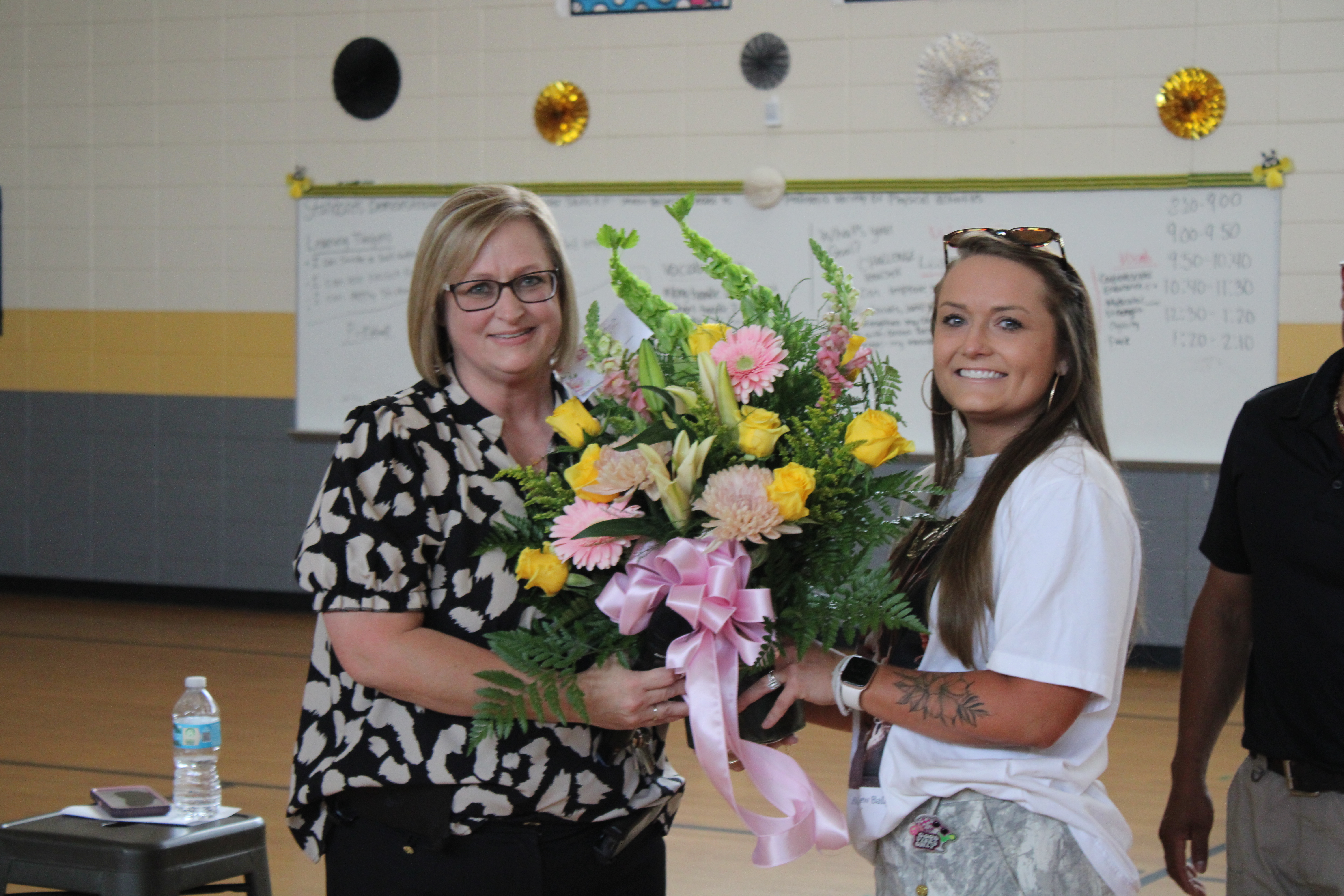 Two women holding a floral bouquet with pink ribbons. A whiteboard and a person in a black shirt are in the background.