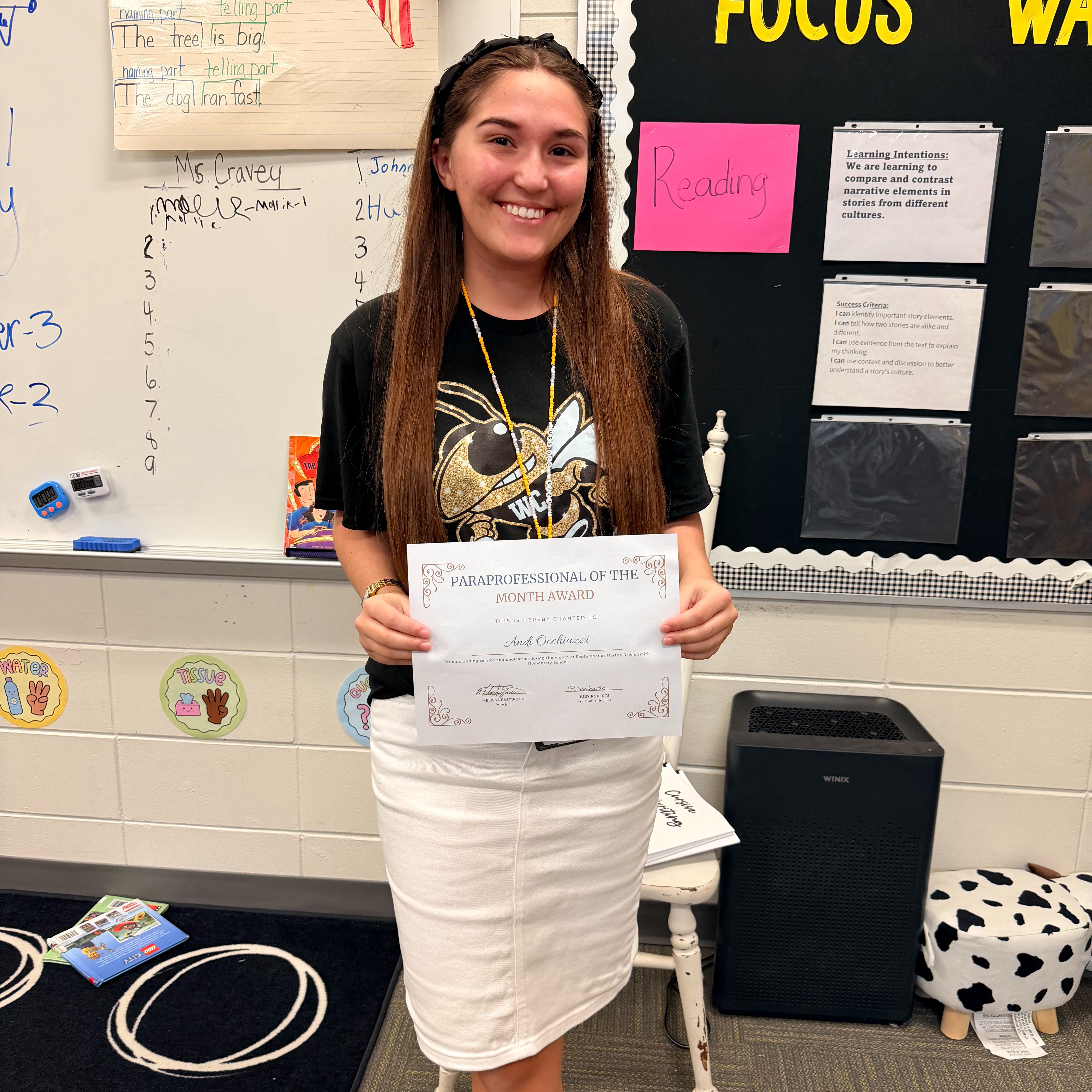 Woman holding a certificate stands in classroom with whiteboards, bulletin board, and a small stool in the background.