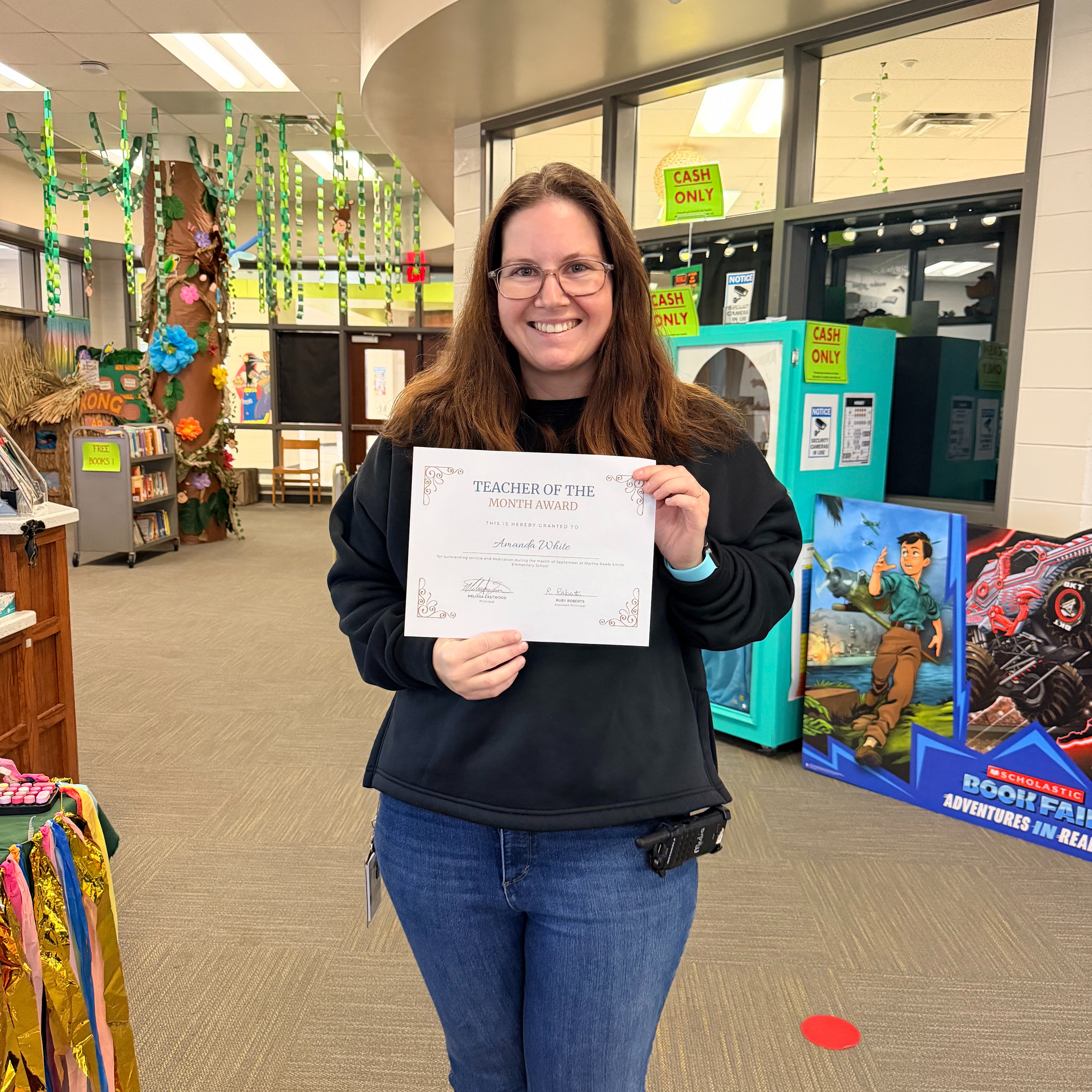 A woman in glasses and a black sweatshirt holds a certificate. Behind her are shelves, a vending machine, and toys.