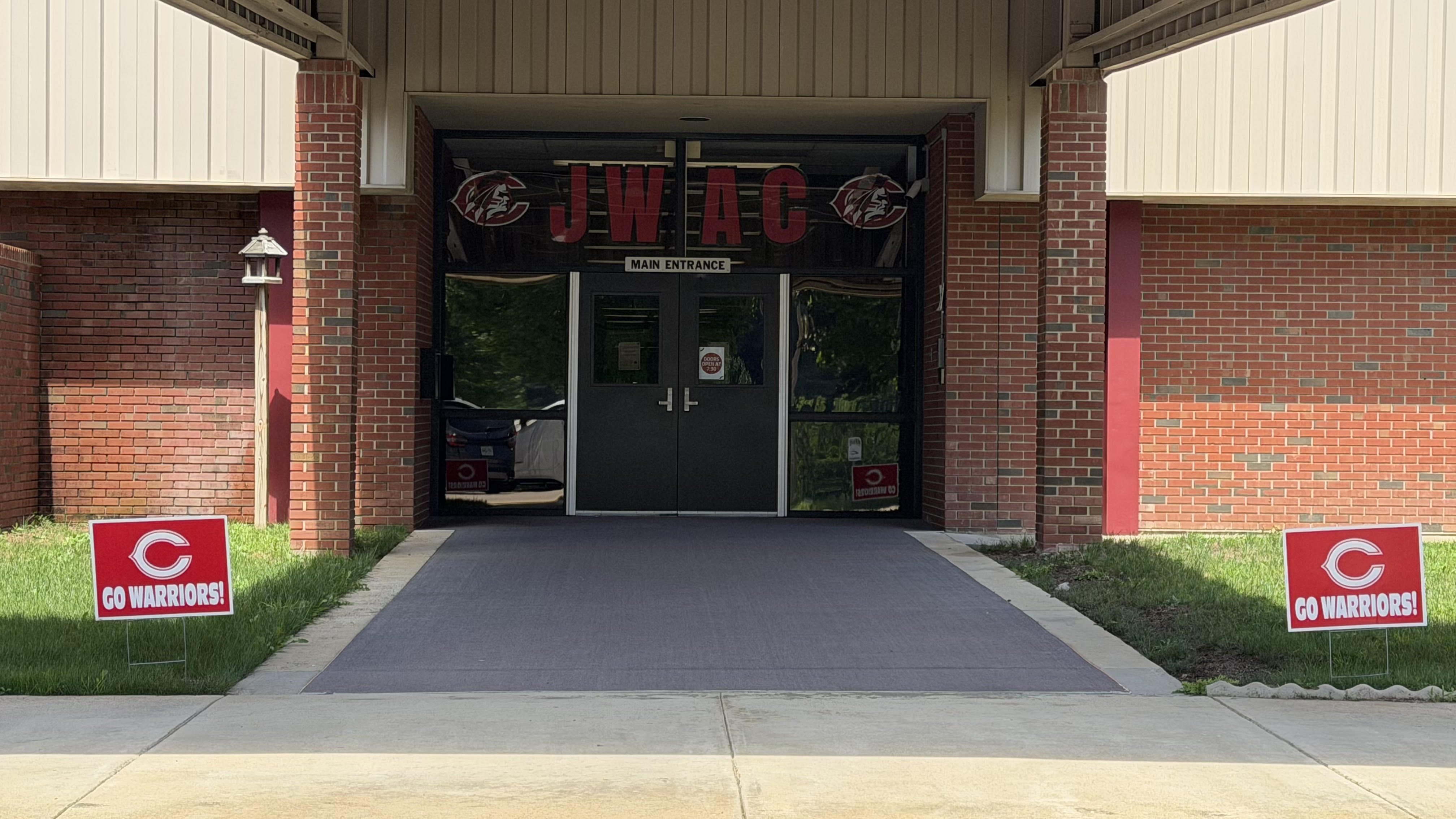 Exterior of a building with brick walls, black double doors, and two red signs with white text on the grass.
