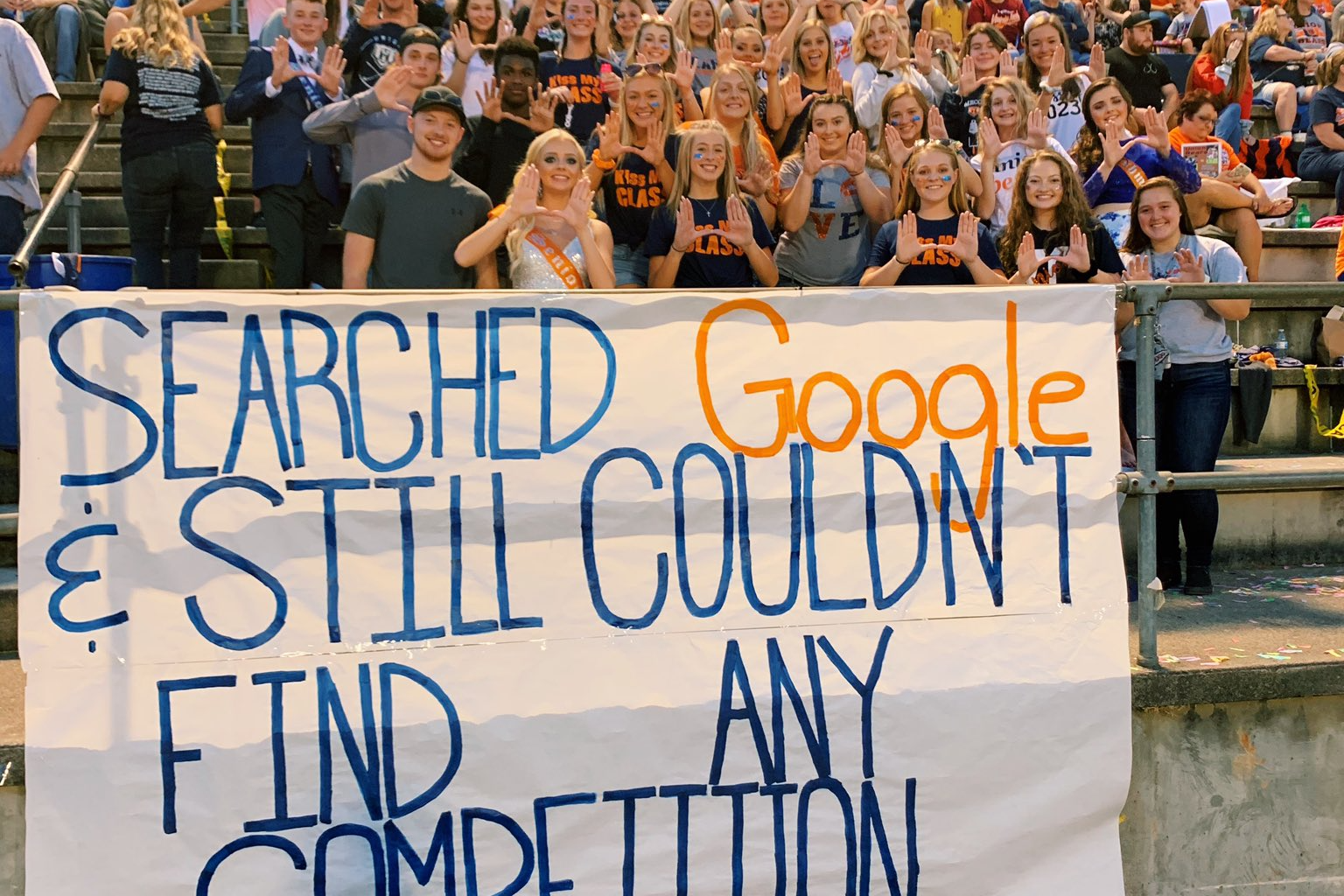 Male and female high school students holding up the U symbol with their hands standing behind a sign that says "Search Google & still couldn't find any competition."