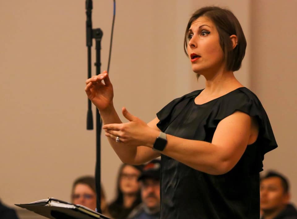 Choral Director Kelly Sorah in a black dress standing at a podium with her hands raised in a conductor position.