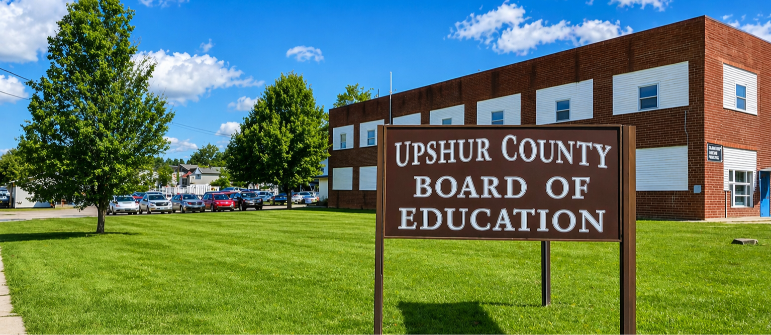 A bright summer day at the Upshur County Board of Education building, where lush green lawns, leafy trees, and a vivid blue sky frame the prominent campus sign in crisp, high-definition detail.